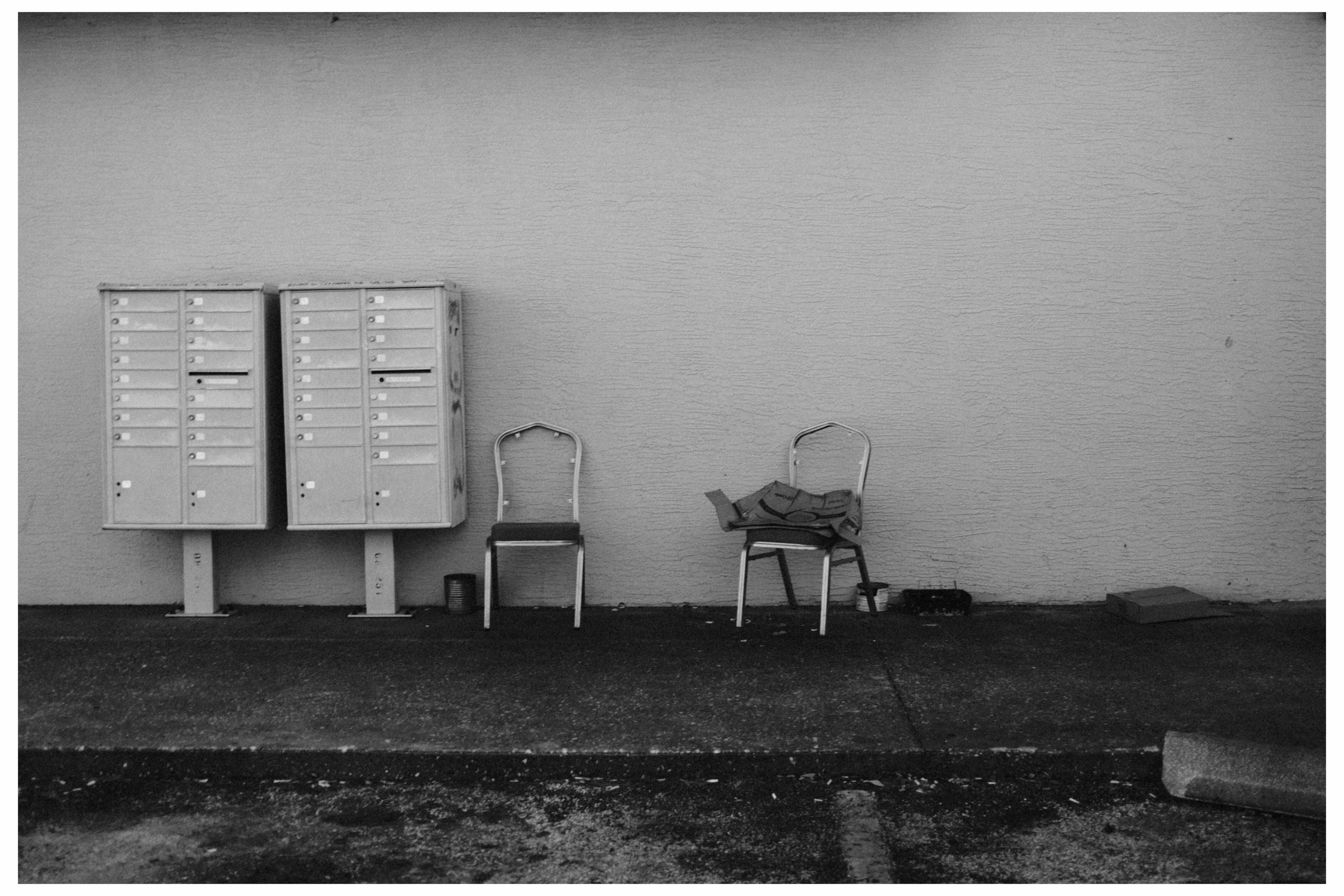 photograph of old chairs with open backs next to two banks of locked mailboxes and in front of a wall