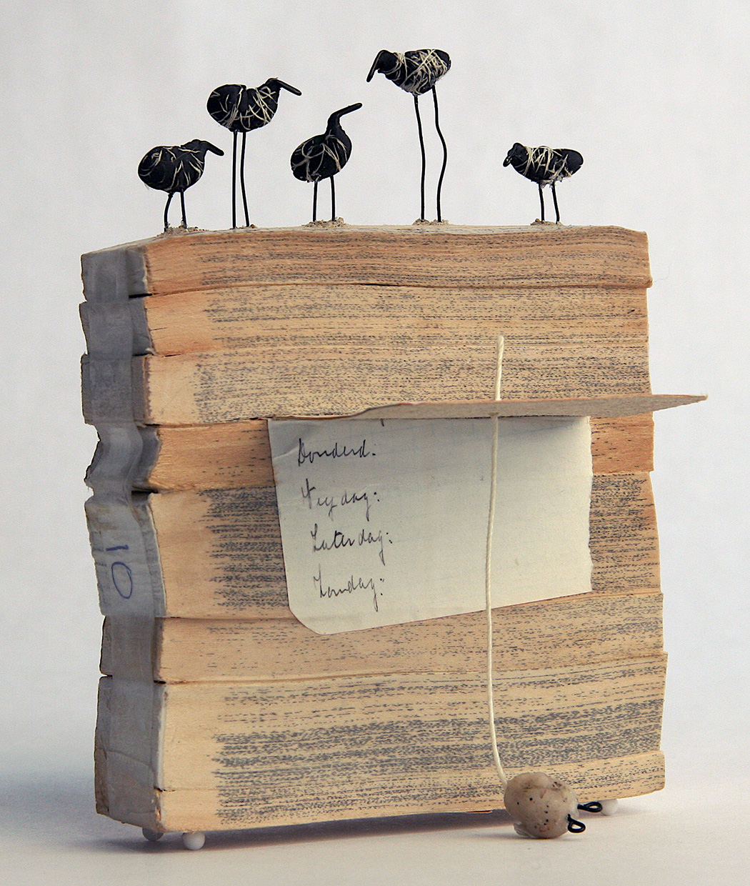 sculpture of a slice of stacked books with five tiny birds on top and on
        			the side it has a folded piece of cardstock with writing on it that 
        			sticks out like a drop-leaf table
        			with a string and little weight attached to it